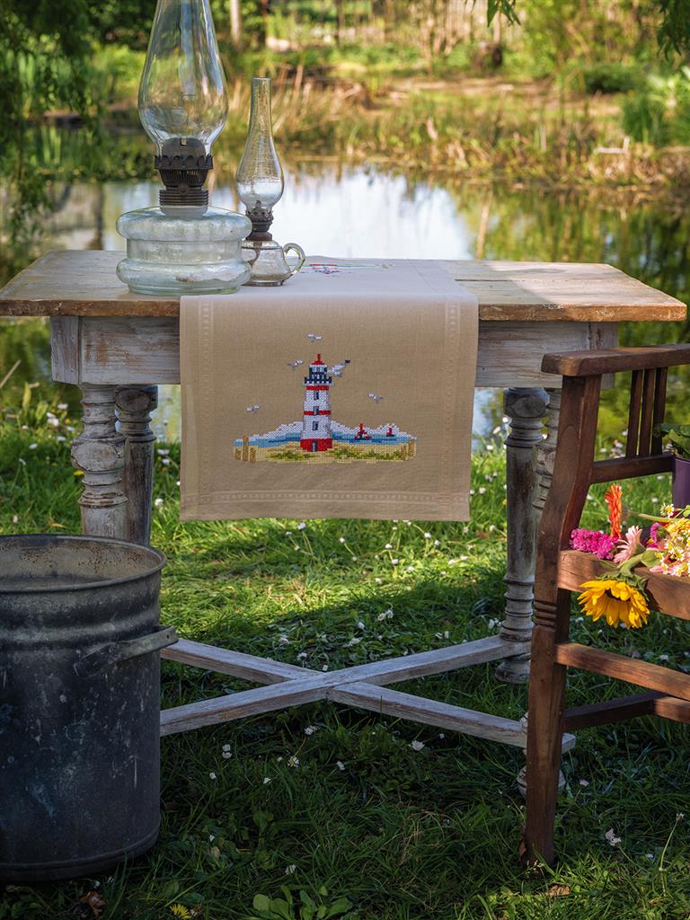 Kit de punto de cruz para crear un camino de mesa de Vervaco con diseño de un faro en la costa de color rojo y blanco. Incluye tela premarcada con dobladillo, hilos DMC y patrón detallado