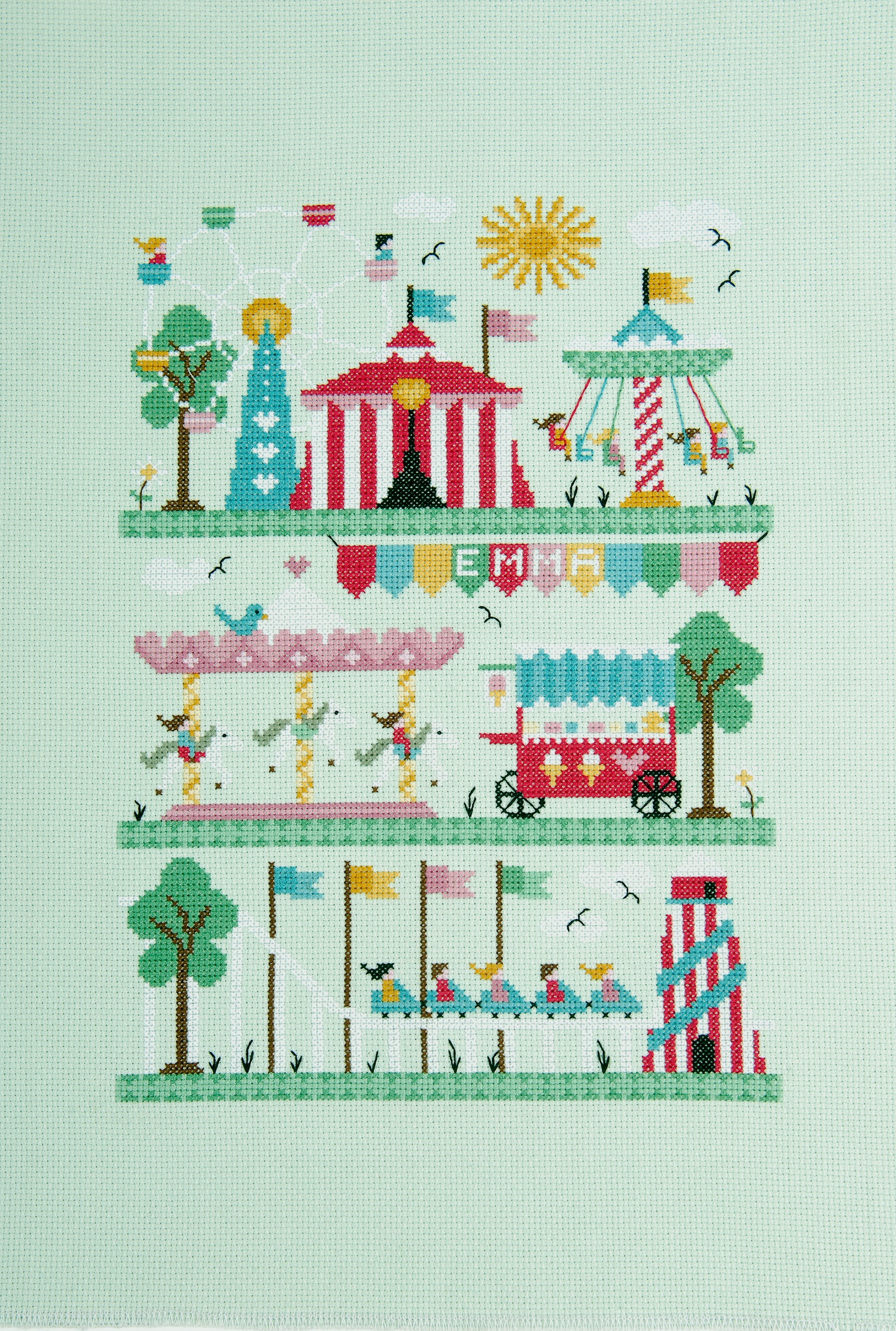 Kit de punto de cruz DMC “Feria” de Faye Walsh con diseño de feria colorida, carruseles y noria bordado en tela Aida 14 ct.