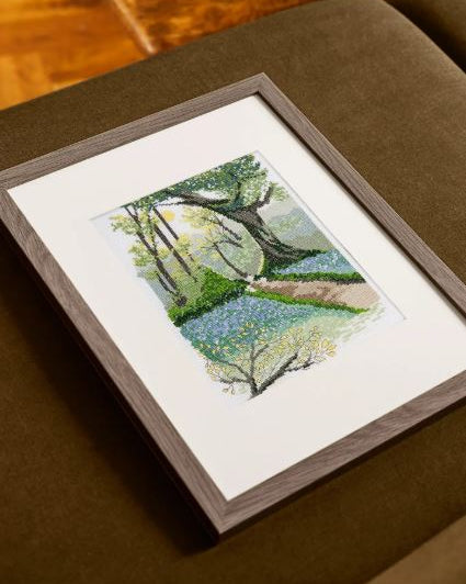 Kit de punto de cruz DMC Paisaje Primaveral con bosque florido, camino y flores en tonos suaves sobre tela Aida blanca.