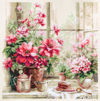 Kit de punto de cruz Magic Needle Morning in Rosy Tones con flores rosadas y escena luminosa junto a una ventana bordada en tela Aida marfil.