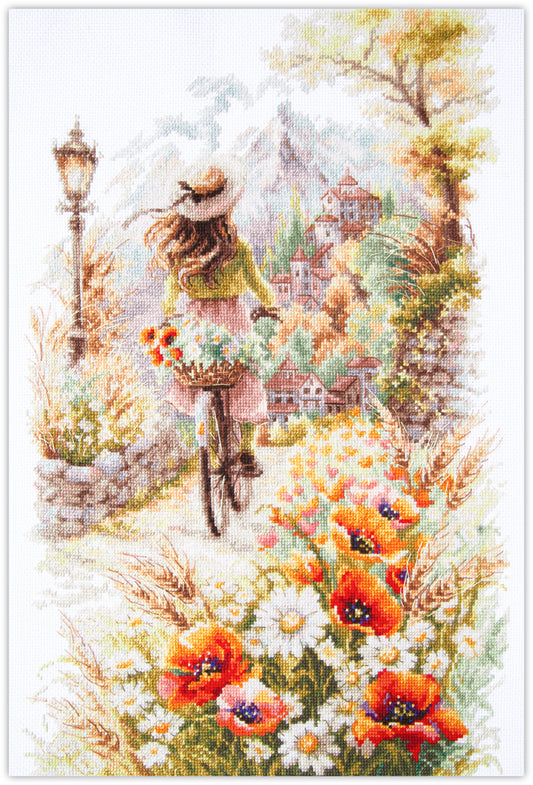 Kit punto de cruz Para Ser Feliz Magic Needle con joven en bicicleta, cesta de flores y campo de amapolas frente a un pueblo entre montañas.